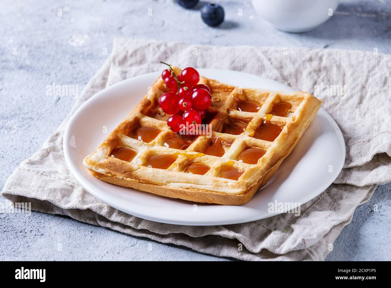 Frühstück mit hausgemachten quadratischen belgischen Waffeln mit frischen reifen Beeren Rote Johannisbeere und Karamellsauce auf weißem Teller mit Krug Von Milch und Textil napki Stockfoto