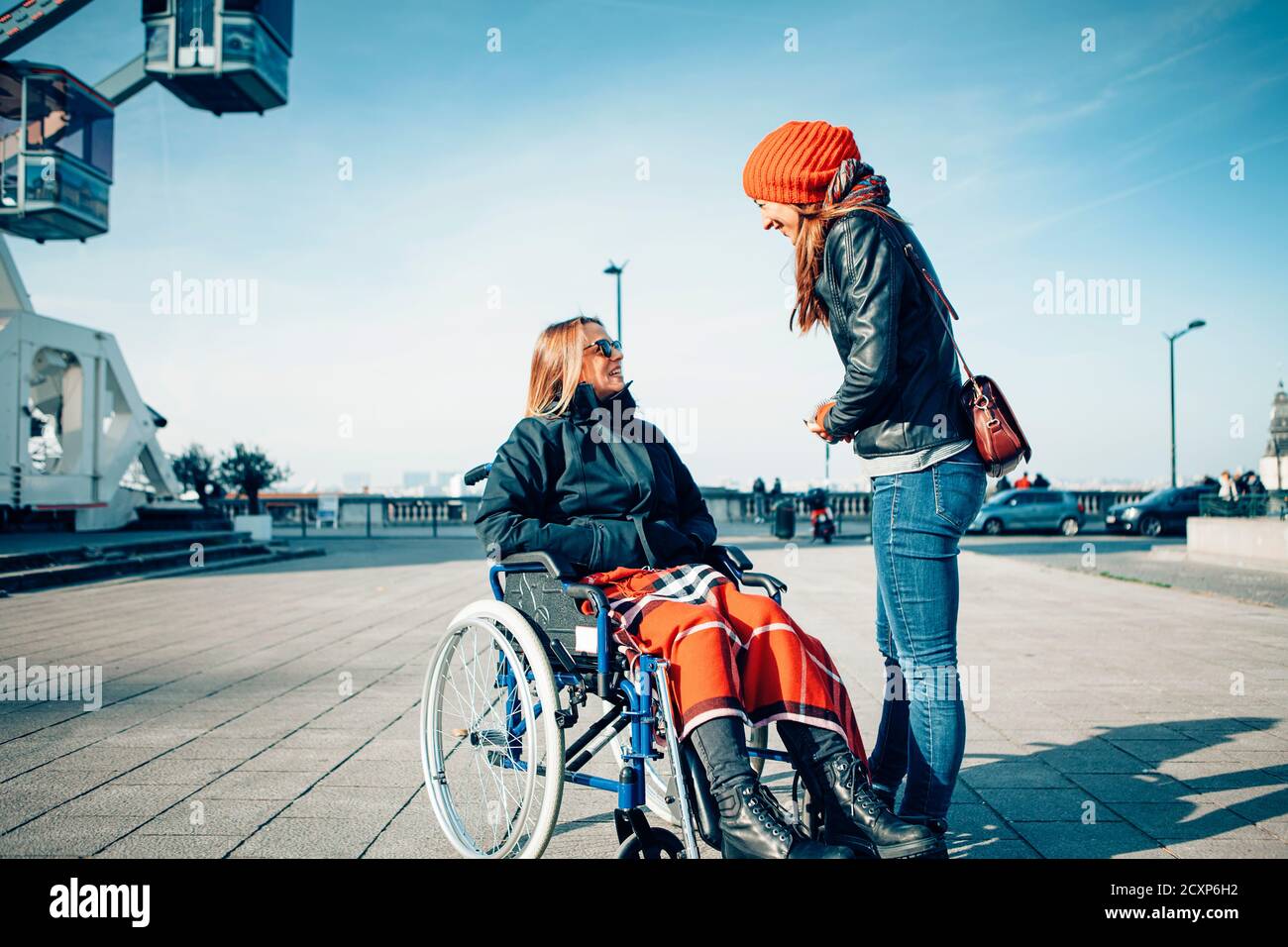 Zwei lächelnde Frauen sprechen und verbringen Zeit zusammen draußen in Die Stadt - sitzt auf einem Rollstuhl und lebt ein Behinderung mit positiver Einstellung Konz Stockfoto