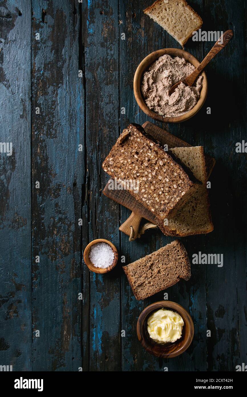 Verschiedene Brote in Scheiben hausgemachtes Roggenbrot Vollkorn und Samen für das Frühstück mit Olivenholz Schüsseln mit Butter, Salz, Leber Paste über alten dunklen Woode Stockfoto