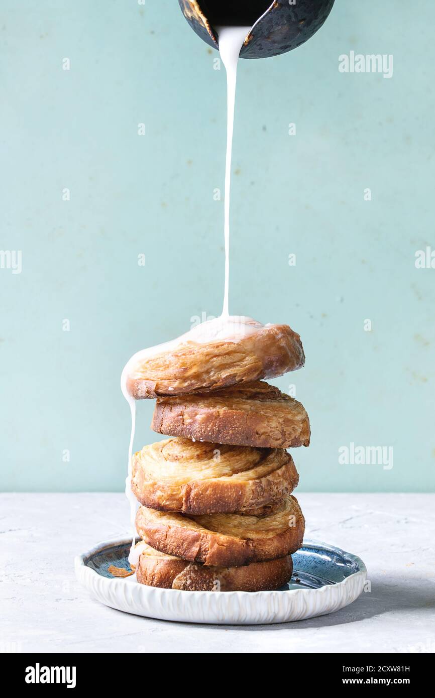 Stapel von hausgemachten verglaste Blätterteig Zimtschnecken mit Vanillesauce und Rosinen auf blauen Platte über Grau grün Hintergrund. Gießen Glasur aus der Kanne. Pin-up Stockfoto