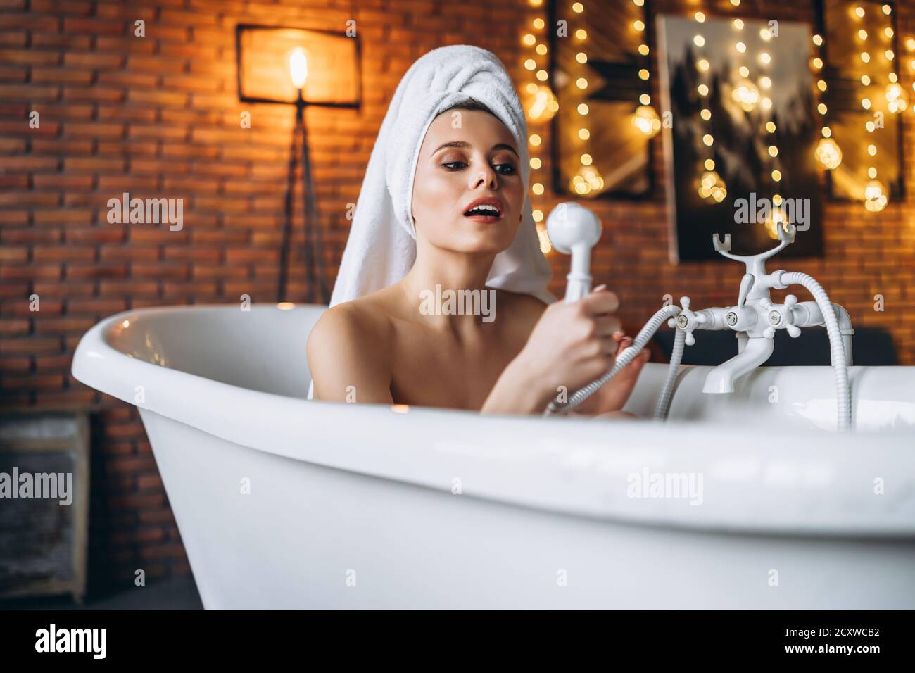 Eine schöne junge Frau posiert im Studio. Frau liegt in einem weißen Bad mit einem Handtuch auf dem Kopf. Eine rote Backsteinmauer mit gelben Girlanden auf dem Backgroun Stockfoto