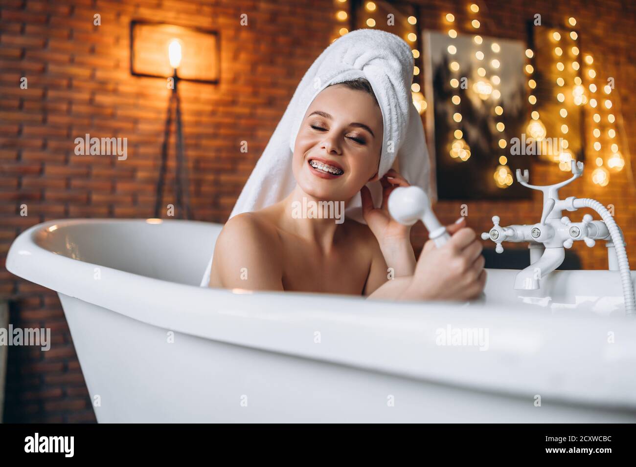 Eine schöne junge Frau posiert im Studio. Frau liegt in einem weißen Bad mit einem Handtuch auf dem Kopf. Eine rote Backsteinmauer mit gelben Girlanden auf dem Backgroun Stockfoto