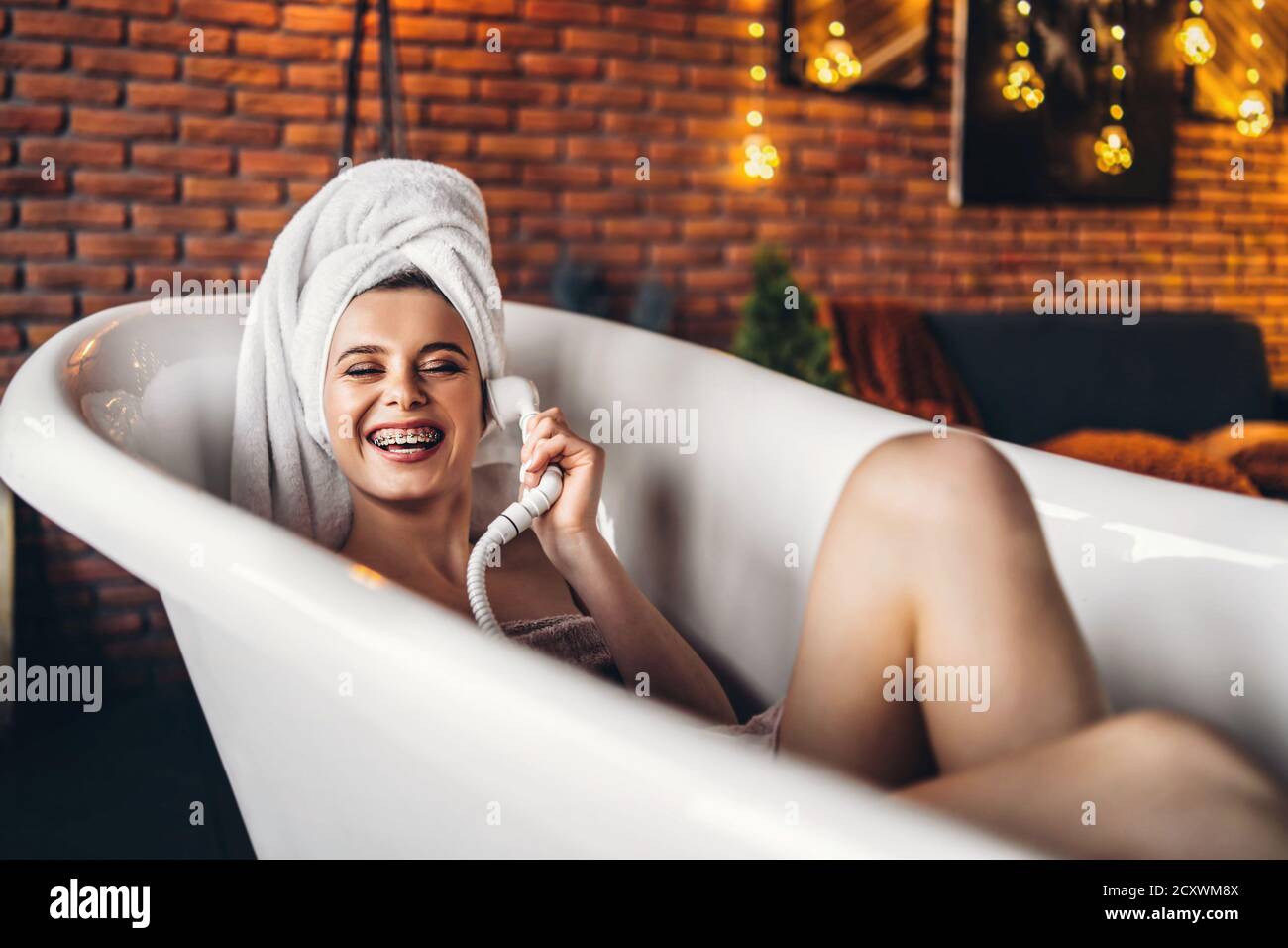 Eine schöne junge Frau posiert im Studio. Frau liegt in einem weißen Bad mit einem Handtuch auf dem Kopf. Eine rote Backsteinmauer mit gelben Girlanden auf dem Hintergrund Stockfoto