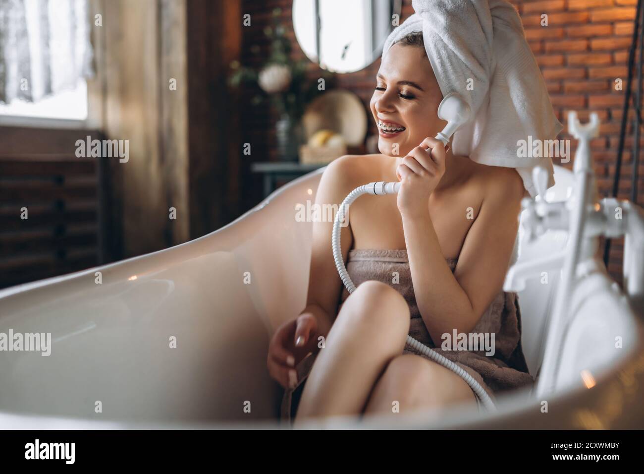 Eine schöne junge Frau posiert im Studio. Frau liegt in einem weißen Bad mit einem Handtuch auf dem Kopf. Eine rote Backsteinmauer mit gelben Girlanden auf dem Hintergrund Stockfoto