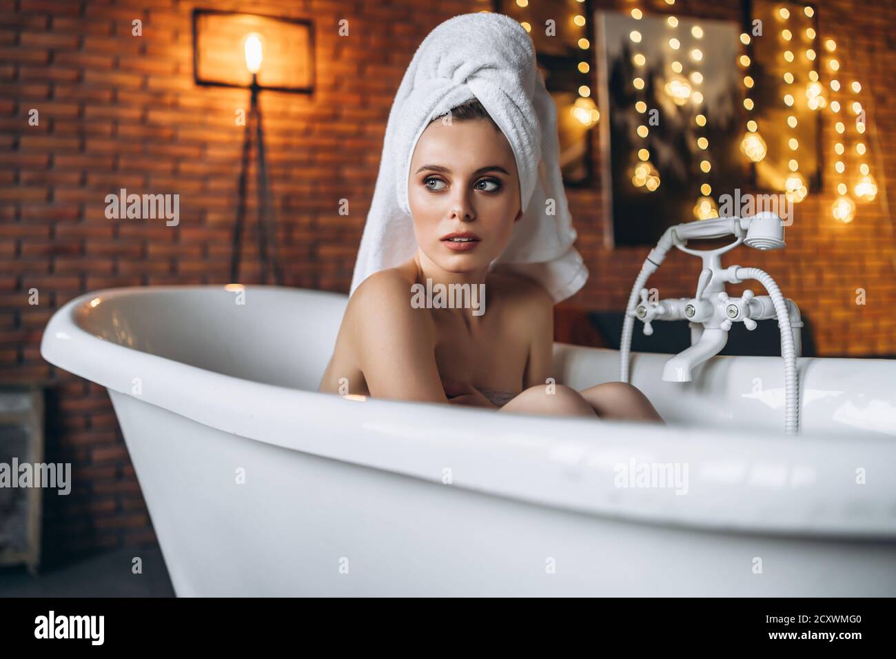 Eine schöne junge Frau posiert im Studio. Frau liegt in einem weißen Bad mit einem Handtuch auf dem Kopf. Eine rote Backsteinmauer mit gelben Girlanden auf dem Hintergrund Stockfoto