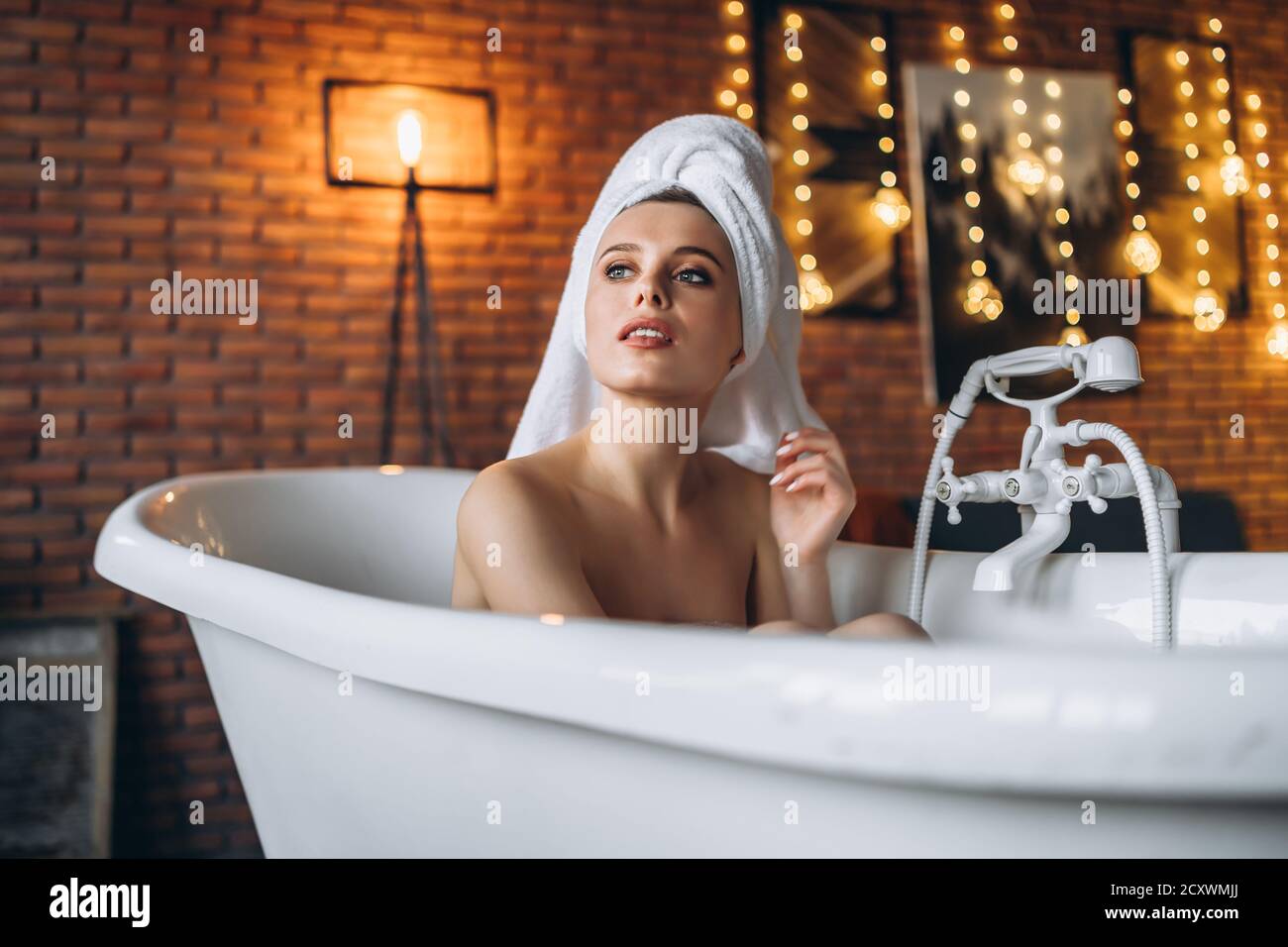 Eine schöne junge Frau posiert im Studio. Frau liegt in einem weißen Bad mit einem Handtuch auf dem Kopf. Eine rote Backsteinmauer mit gelben Girlanden auf dem Hintergrund Stockfoto