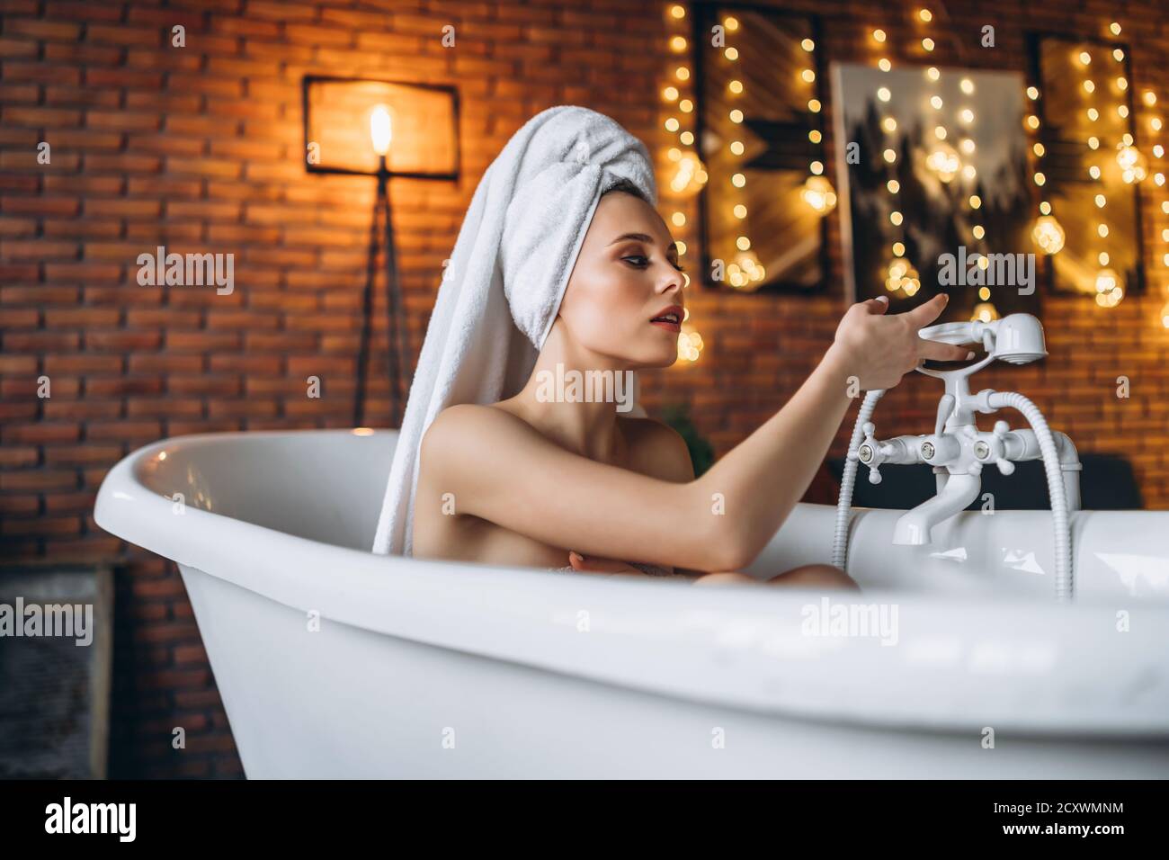 Eine schöne junge Frau posiert im Studio. Frau liegt in einem weißen Bad mit einem Handtuch auf dem Kopf. Eine rote Backsteinmauer mit gelben Girlanden auf dem Hintergrund Stockfoto