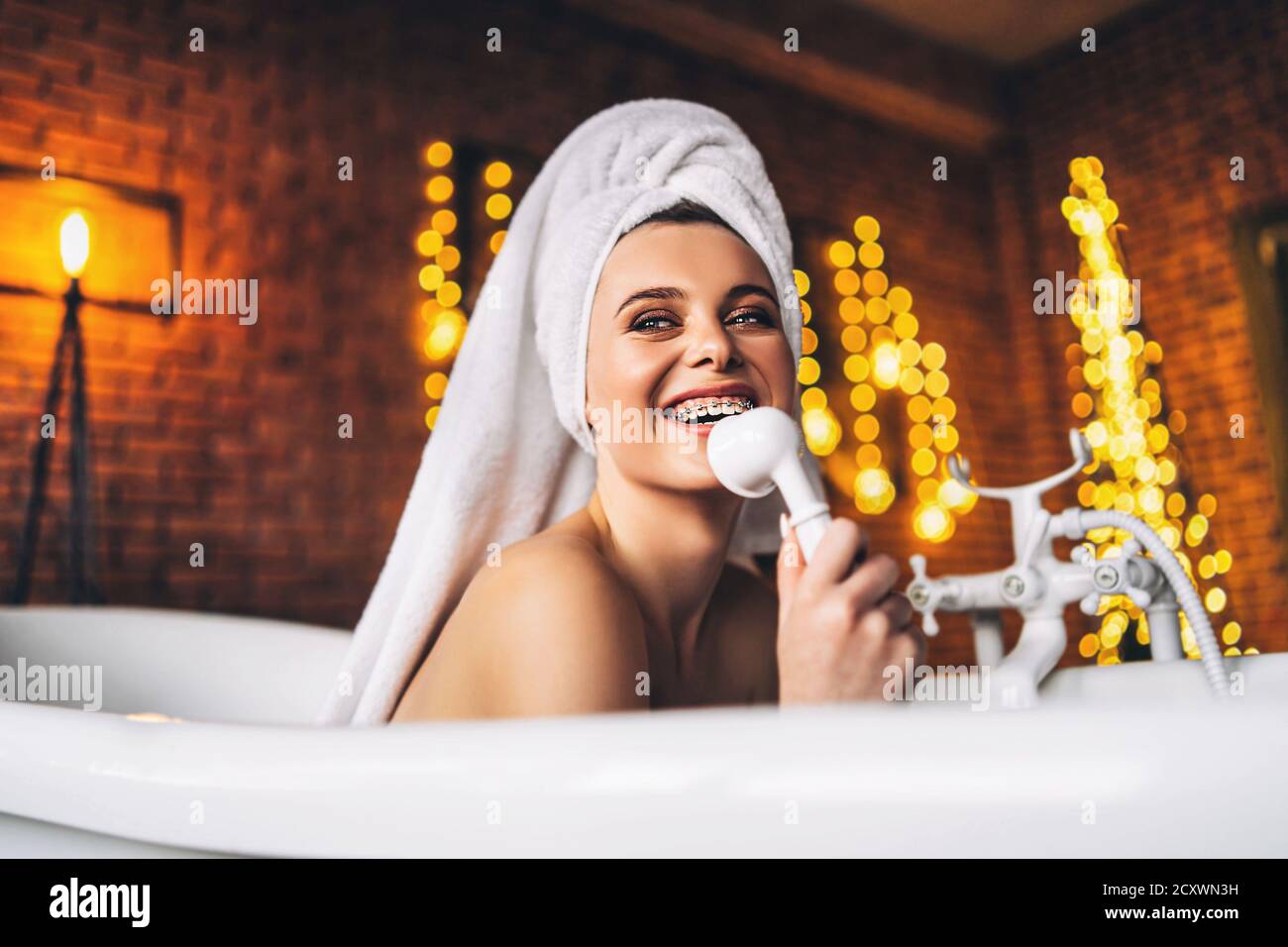 Eine schöne junge Frau posiert im Studio. Frau liegt in einem weißen Bad mit einem Handtuch auf dem Kopf. Eine rote Backsteinmauer mit gelben Girlanden auf dem Hintergrund Stockfoto