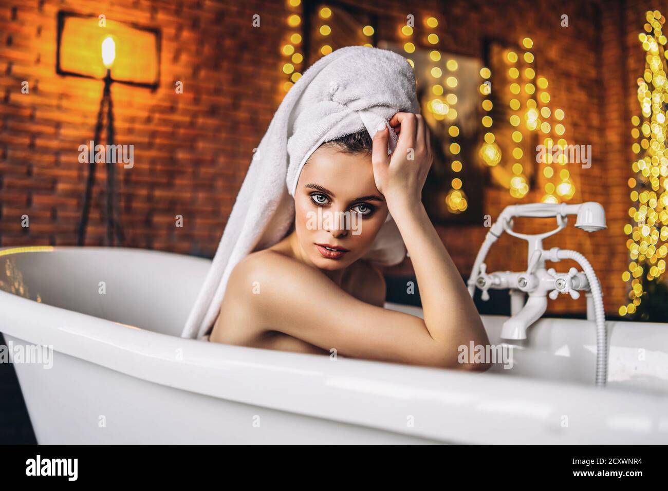 Eine schöne junge Frau posiert im Studio. Frau liegt in einem weißen Bad mit einem Handtuch auf dem Kopf. Eine rote Backsteinmauer mit gelben Girlanden auf dem Hintergrund Stockfoto