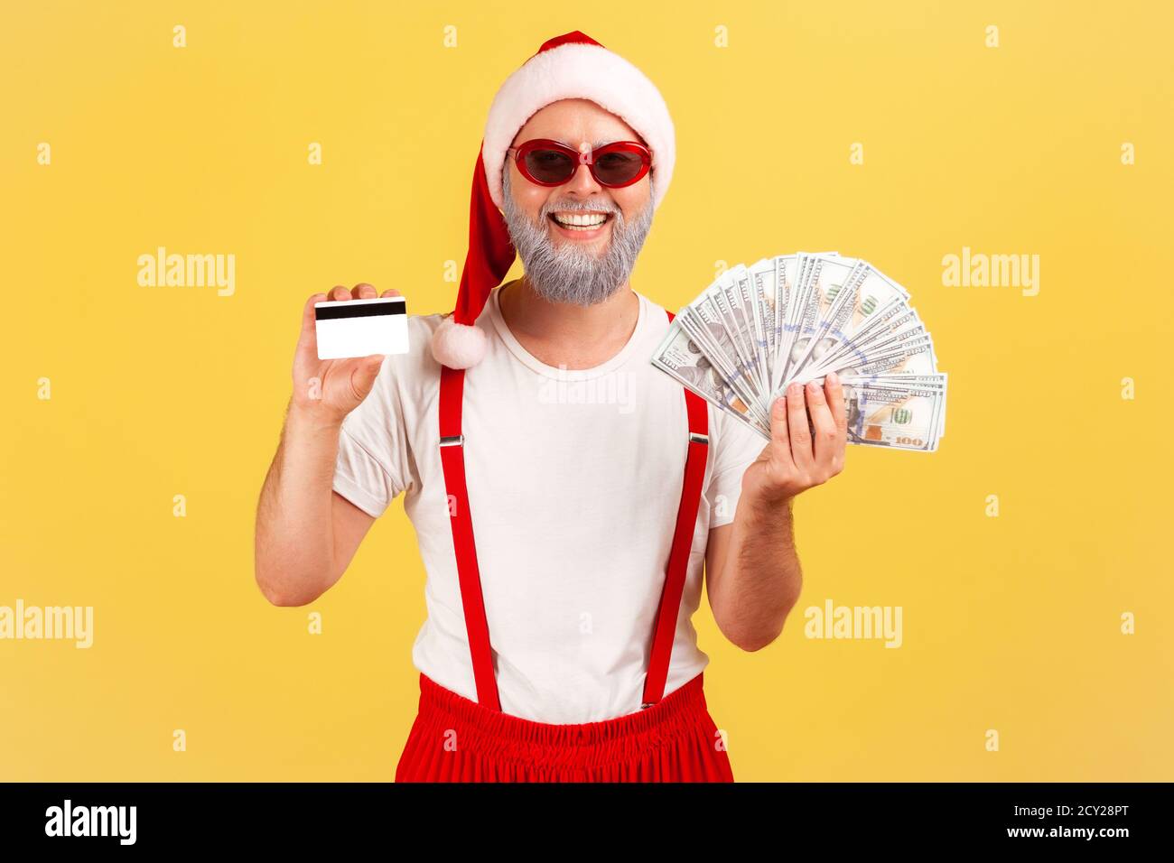 Aufgeregter, positiver graubärtiger Mann mit weihnachtsmann-Hut und Sonnenbrille, der Kreditkarten- und Dollarnoten hält und mit toothy Lächeln auf die Kamera schaut. Indo Stockfoto