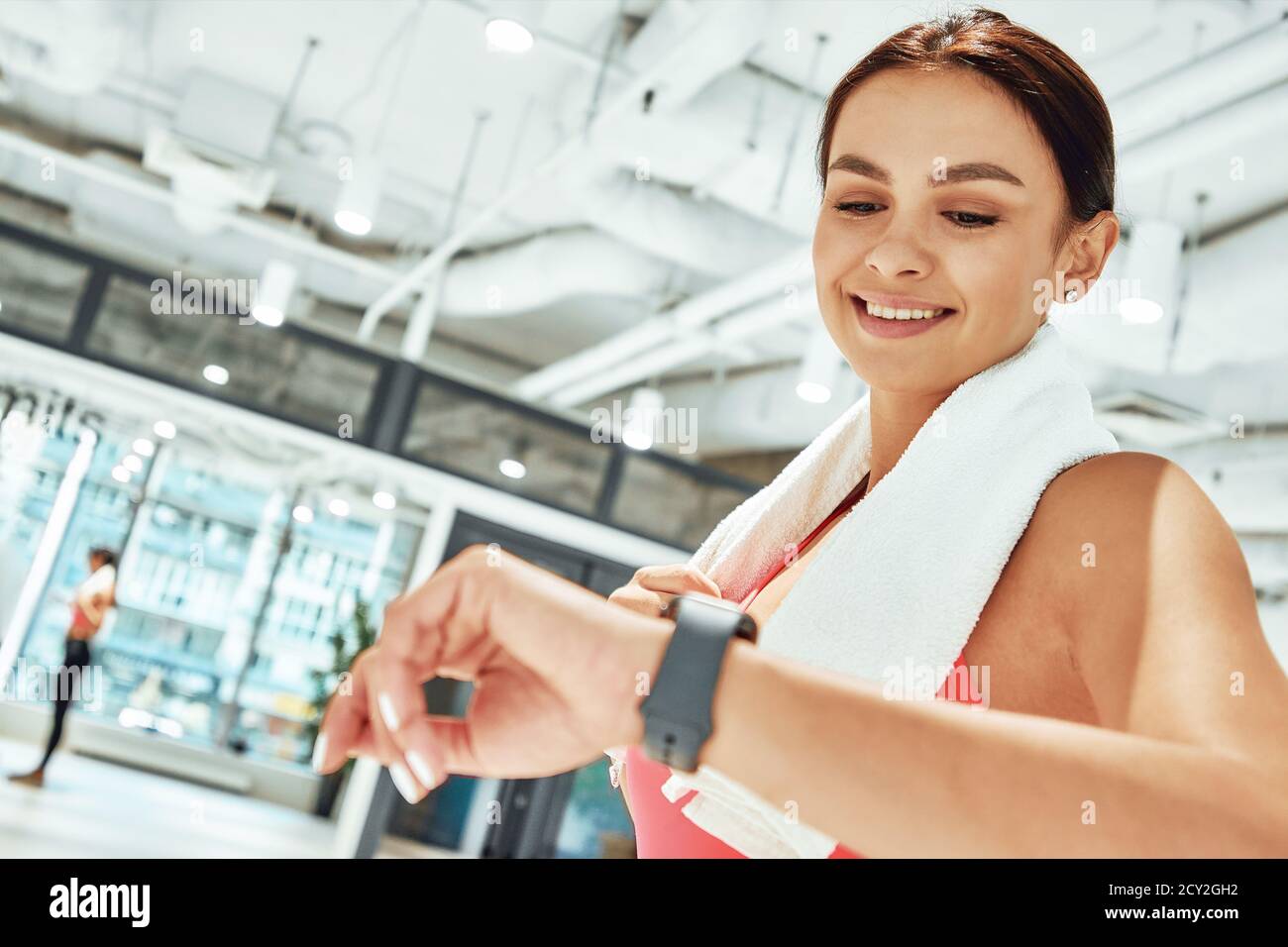 Junge glückliche sportliche Frau mit weißem Handtuch auf den Schultern Blick auf Smartwatch beim Training im Fitnessstudio. Sport, Workout, Wellness und gesundes Lifestyle-Konzept Stockfoto