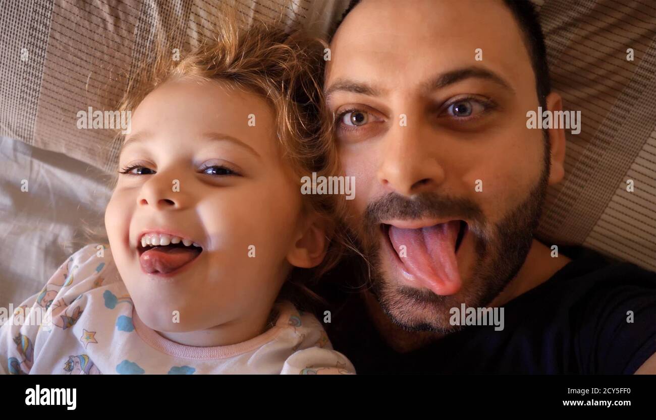 Authentische Nahaufnahme eines Vaters und einer Tochter, die lustige Gesichter machen, auf den Bildschirm für ein Selfie schauen, in einem Bett. Konzept der Familie und emotionale Beziehungen Stockfoto