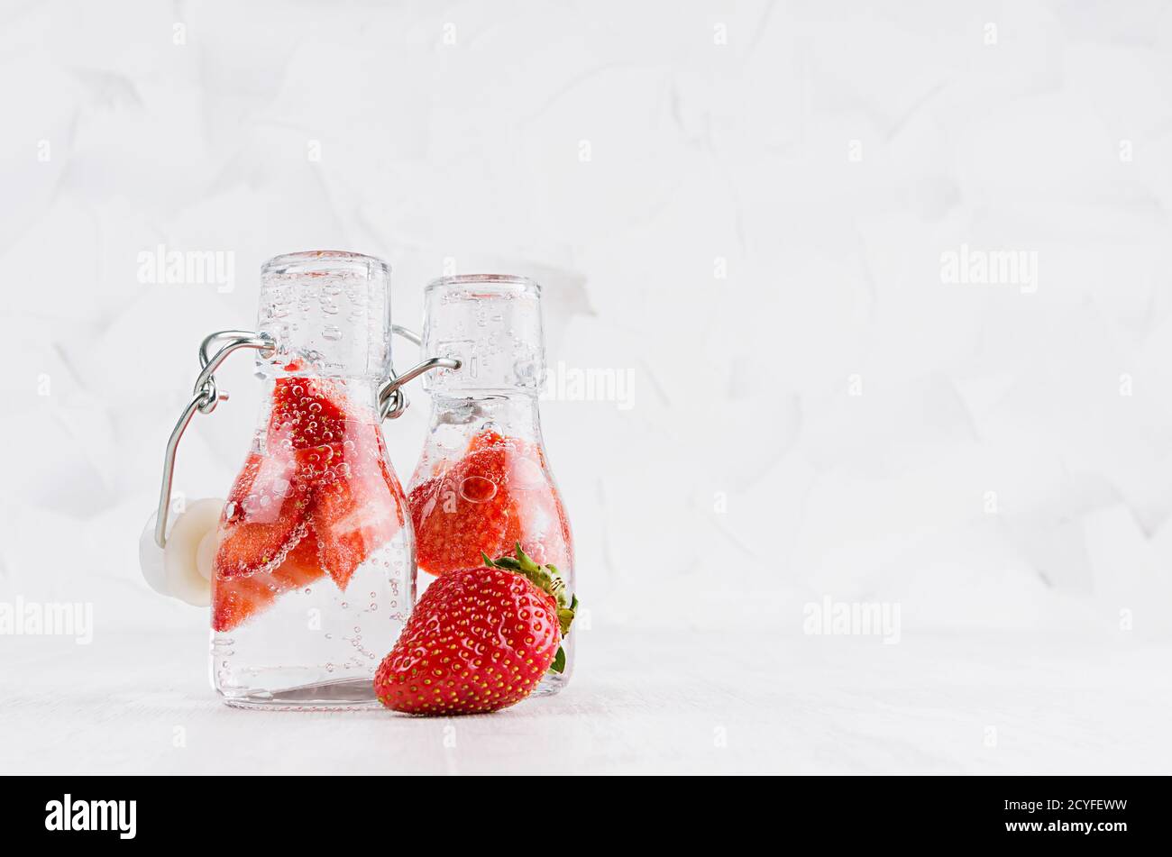 Erfrischende kalte Fruchtgetränke mit roter Erdbeere, helle Blasen in transparenten Yoke-Flaschen im eleganten weißen Interieur auf Holztisch, Kopierraum. Stockfoto