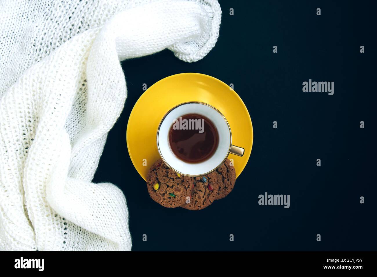 Weiß gestrickt Stoff, Porzellan Gold Tasse mit Kaffee und gelben Teller mit aromatisierten Cookies mit farbigen Tropfen auf schwarzem Hintergrund. Handwerk auf schwarzem Bac Stockfoto