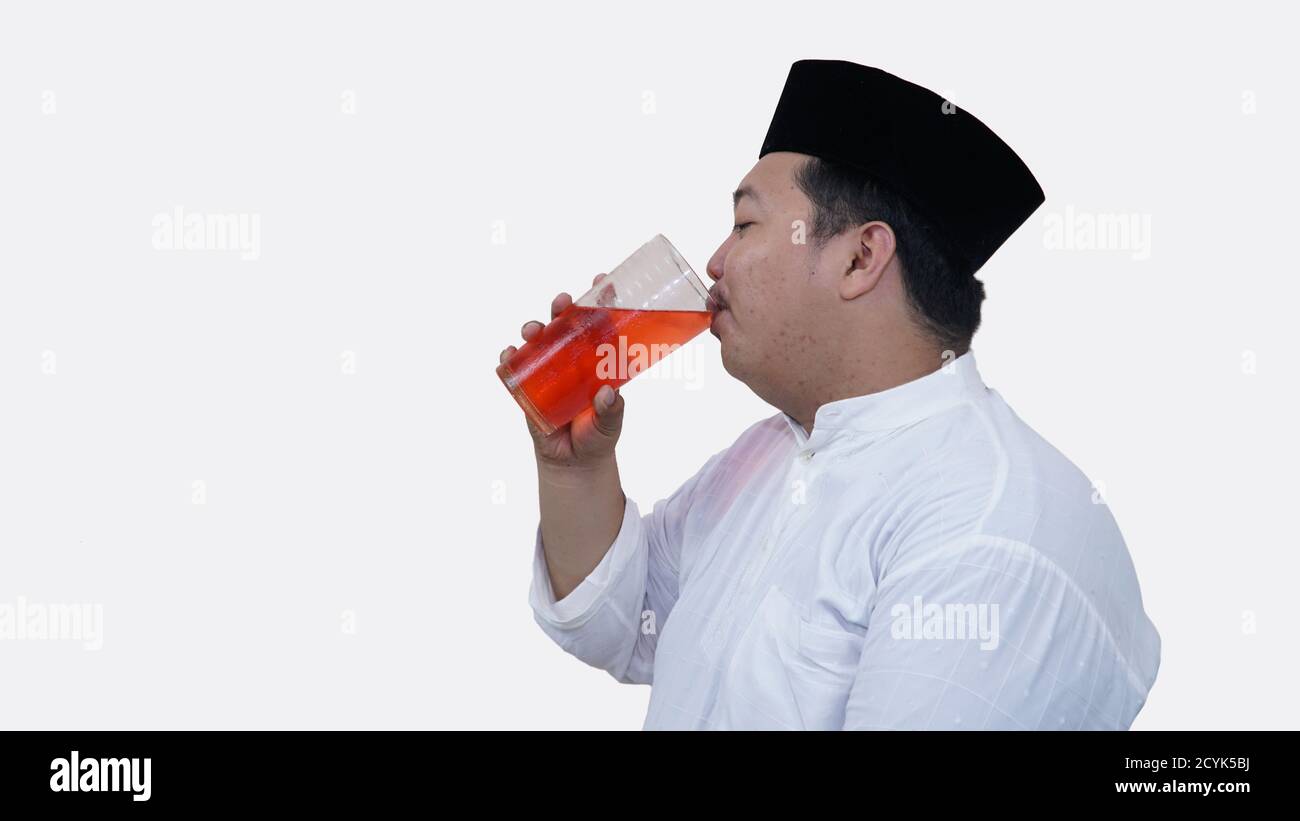 Porträt eines übergewichtigen muslimischen Mannes mit Kopfhaube oder Songkok Trinken Sie ein großes Glas Tee, um das Fasten zu brechen Im Studio drinnen Stockfoto