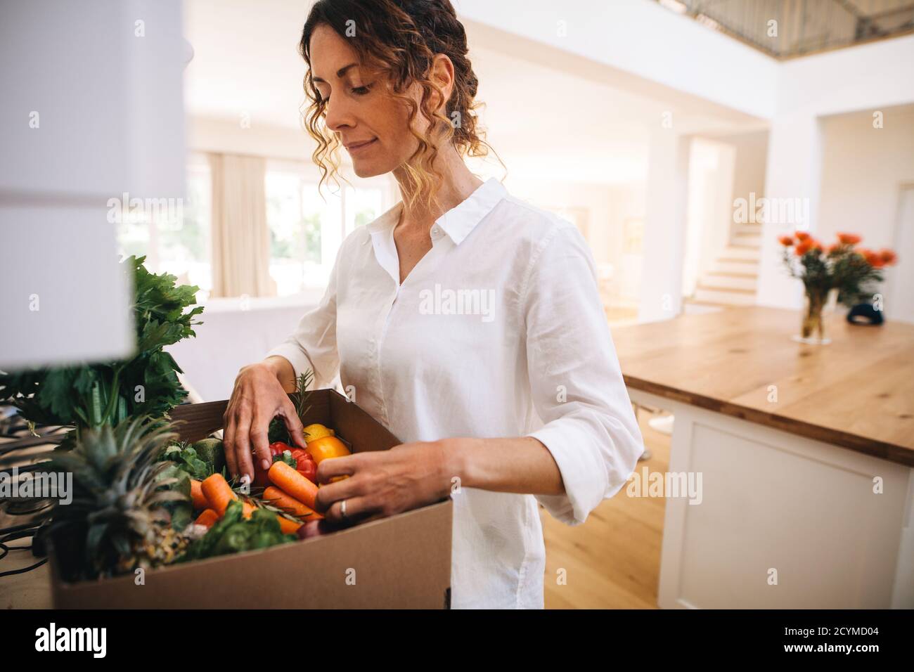 Frau in ihrer Küche mit einer Schachtel voller Bio-Gemüse. Weibliche Überprüfung der frischen Gemüse und Obst in einer Lieferbox. Stockfoto