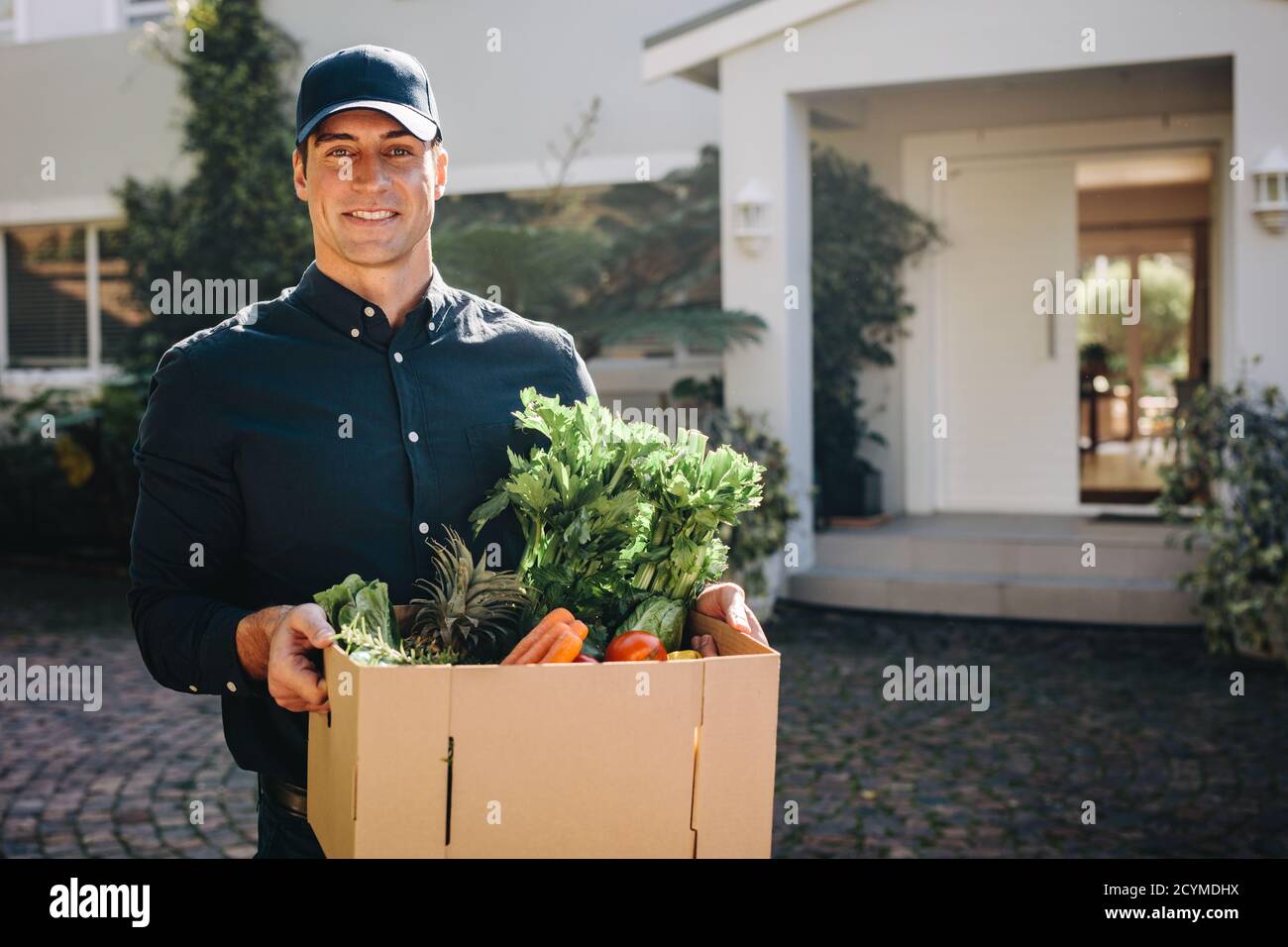 Mann hält eine Schachtel Obst und Gemüse im Freien und schaut auf die Kamera. Lieferperson, die online bestellte Lebensmittel an die Heimatadresse liefert. Stockfoto