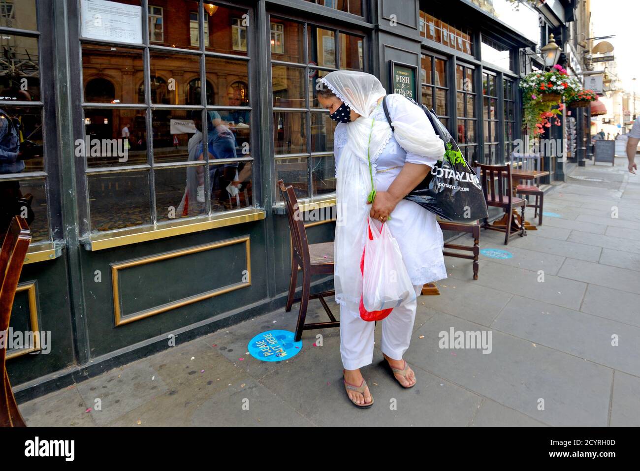 London, England, Großbritannien. Asiatin in Whitehall, trägt eine Gesichtsmask und schaut auf einen 'Bitte warten Sie hier' Aufkleber auf dem Bürgersteig, während der COVID Pandem Stockfoto