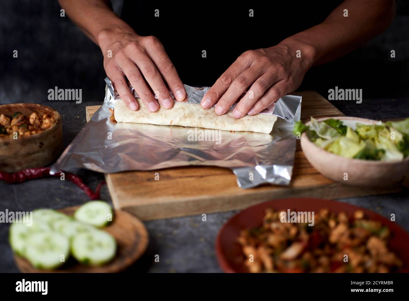 Nahaufnahme eines jungen kaukasischen Mannes, der ein Durum oder einen Burrito zubereitet, mit Hühnerfleisch, das mit verschiedenen Gemüsesorten wie Zwiebeln oder rotem und grünem Pep zubereitet wird Stockfoto
