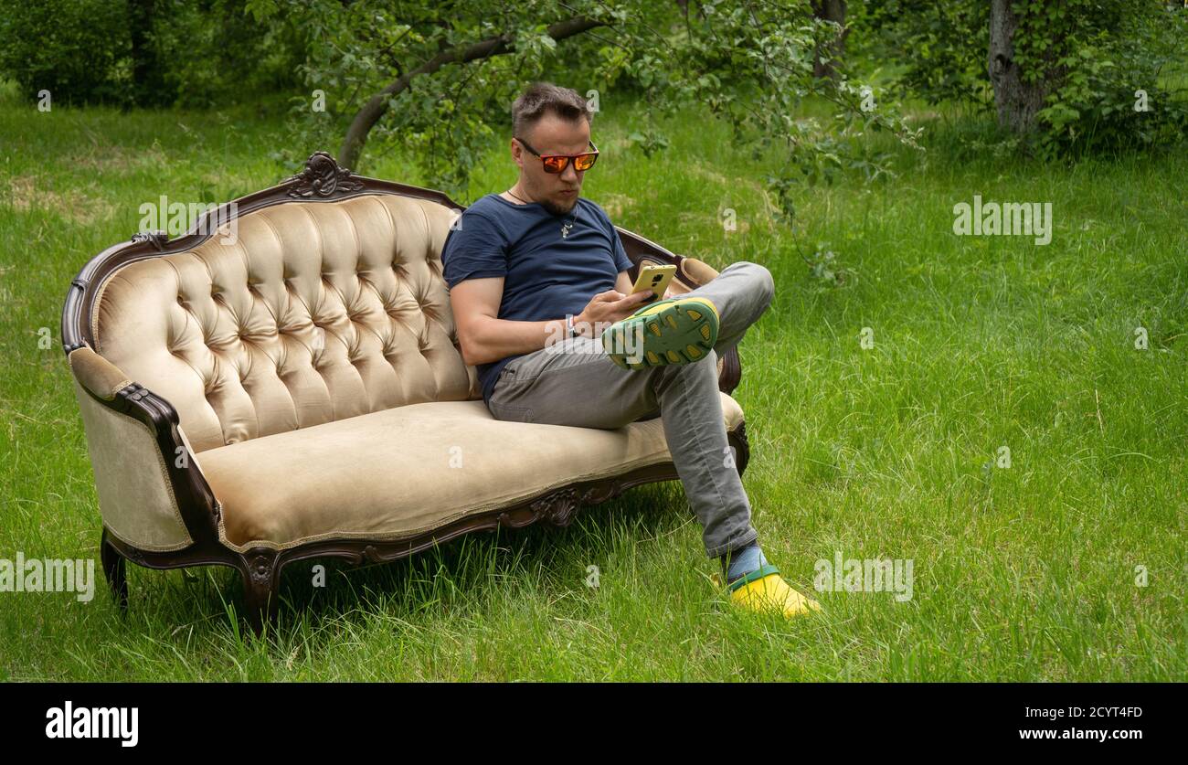 Der Mann mittleren Alters benutzt ein Mobiltelefon, das auf einem hellen Sofa im Garten ruht. Sofa ist auf grünem Gras im Freien. Hochwertige Fotos Stockfoto