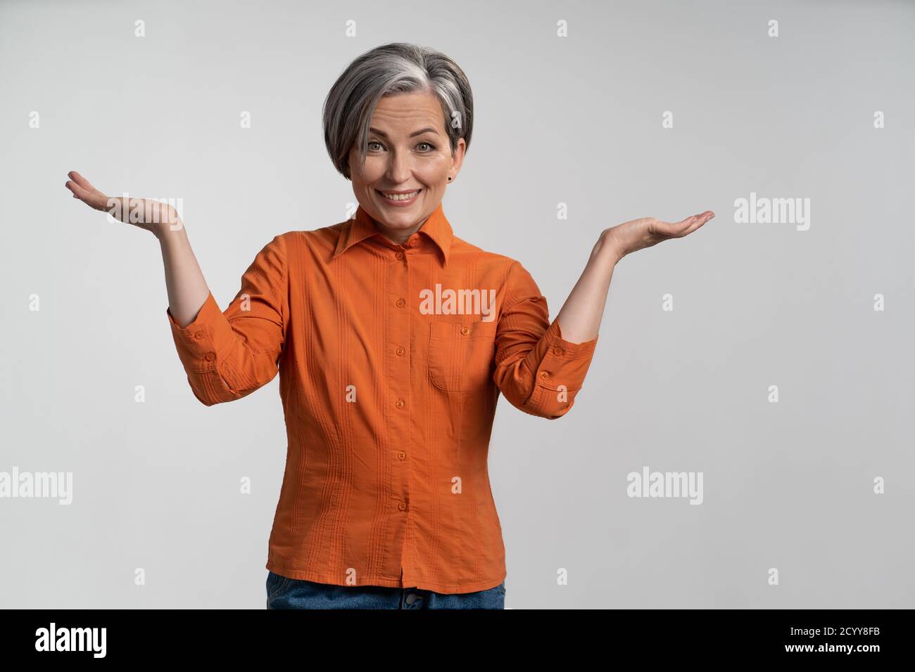 Eine fröhliche Frau mit grauem Haar spreizt ihre gebeugten Arme und lächelt auf die Kamera, während sie an beiden Seiten Kopierflächen zeigt. Erwachsene Frau in orangefarbenem Hemd auf einem Stockfoto