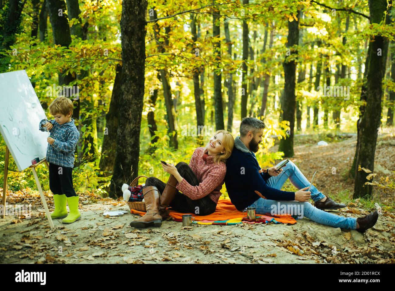 Schöne junge lächelnde Mutter sitzt auf Picknick-Decke mit ihrem entzückenden Mann, während kleine Kind malt mit Bürsten und Zeichnung Staffelei. Glücklich Stockfoto