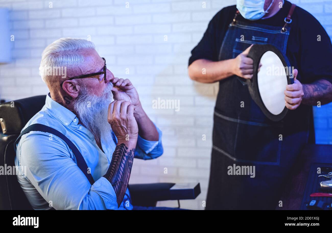 Männliche Friseur schneiden Haare zu Hipster Senior Client während des Tragens Gesicht chirurgische Maske - Junge Friseurin arbeitet in Friseursalon während corona-Virus Stockfoto