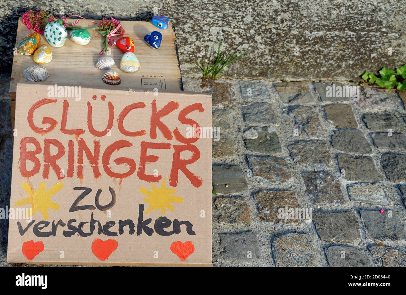 Handbemalte Steine auf dem Bürgersteig mit einem Pappschild mit einem Titel in deutscher Sprache angezeigt: Glück Bringer zu verschenken. Stockfoto