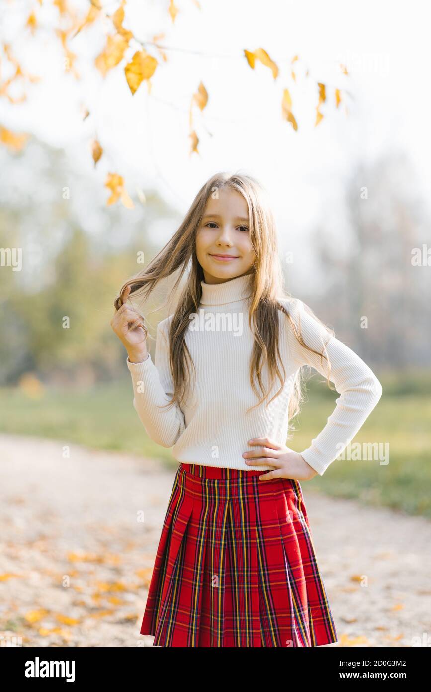 Vorschulkinder kleines Mädchen in einem weißen Pullover und rot karierten Rock mit Herbstblättern im Park. Kindermode. Nettes Baby Mädchen 7-9 Jahre alt Stockfoto