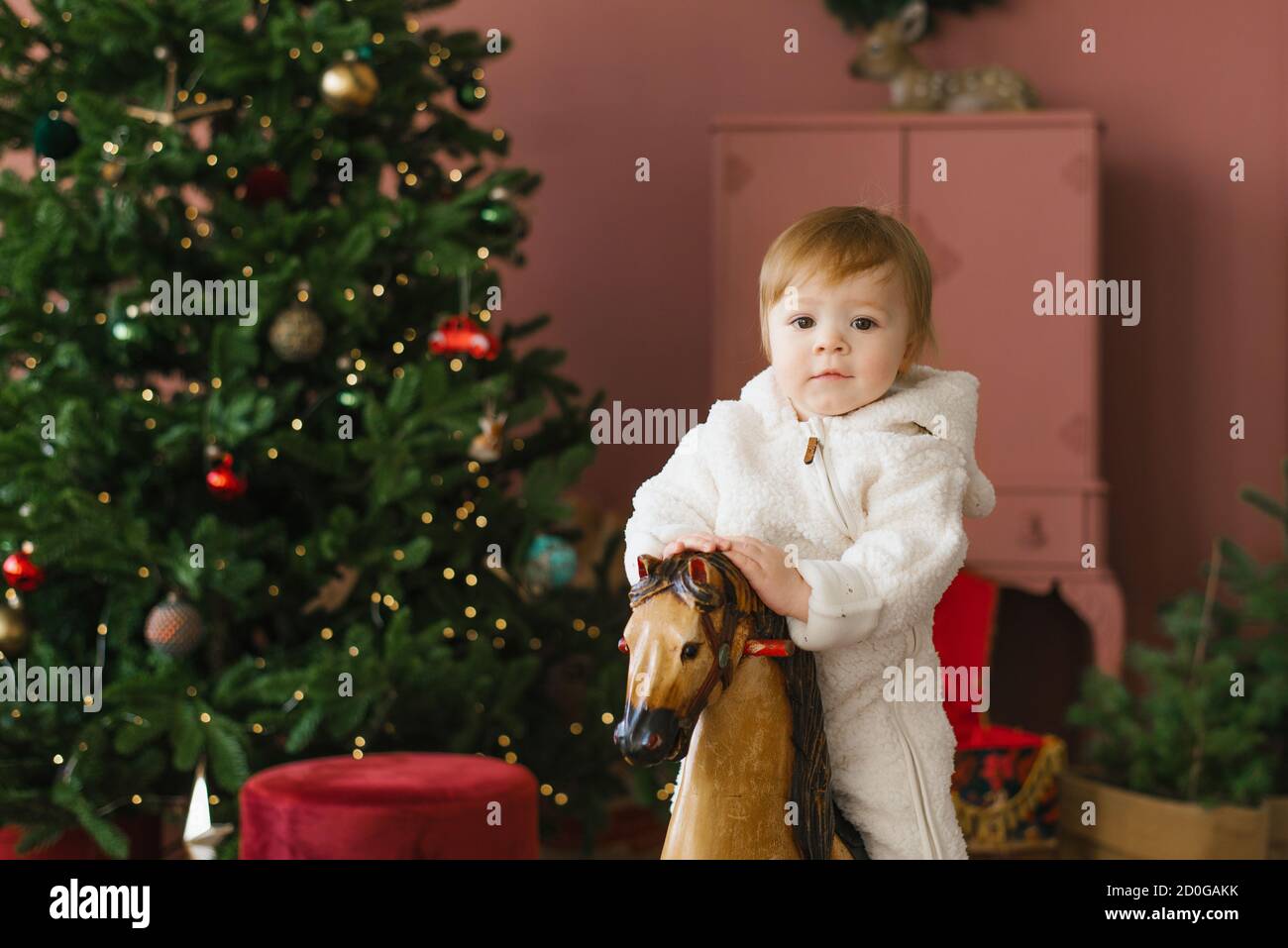 Ein kleines Kind von einem Jahr sitzt auf einem Holzpferd in der Nähe des Weihnachtsbaums Stockfoto