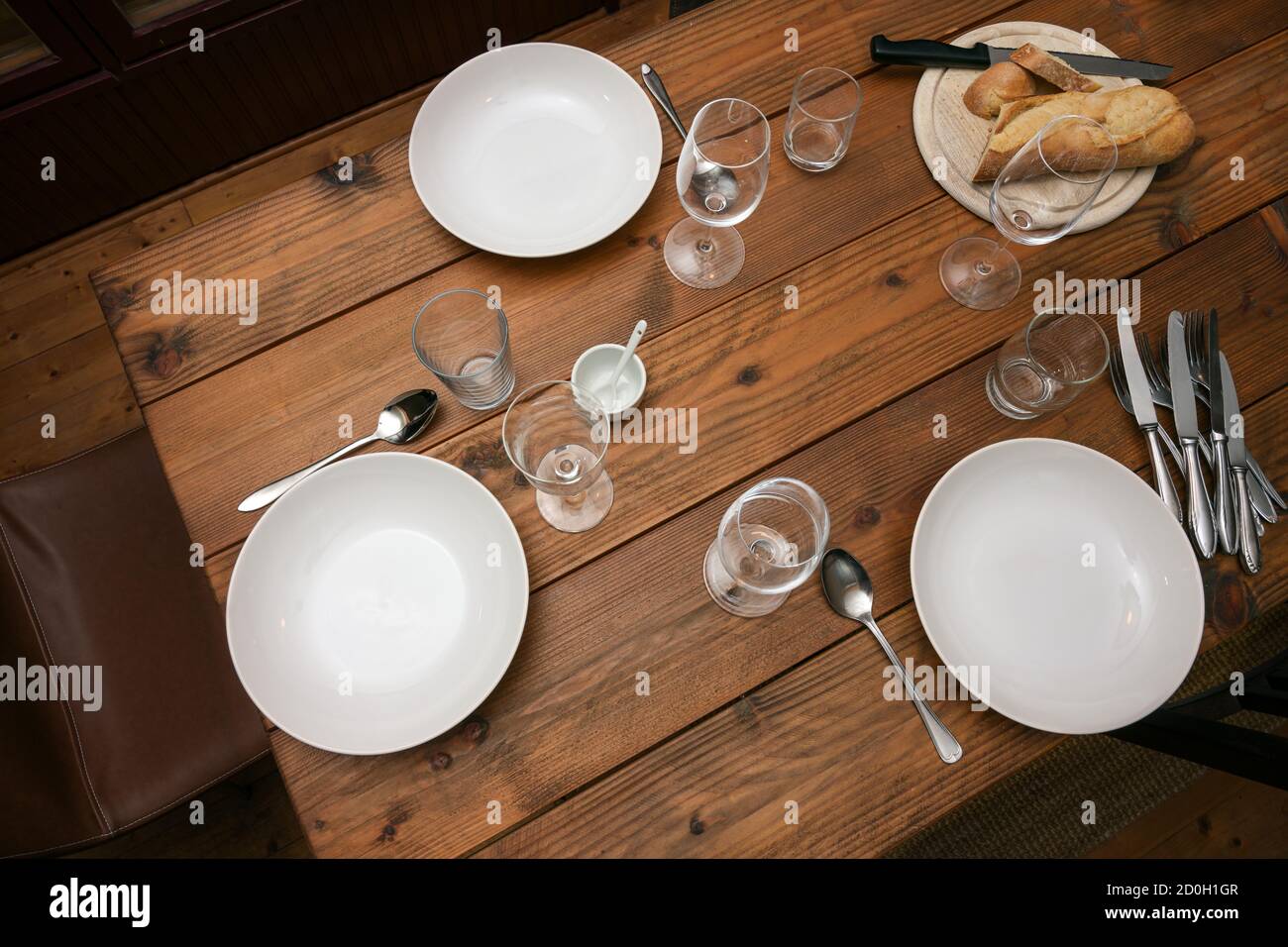 Rustikaler Holztisch halb gelegt mit Tellern, Gläsern, Besteck und Brot für eine informelle Mahlzeit mit Familie oder Freunden, ausgewählte Fokus Stockfoto