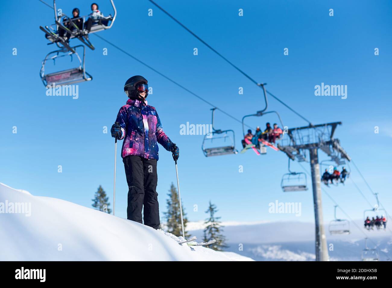 Lächelnde Skifahrerin Frau in Brille und Helm auf ihren Skiern mit Stöcken in der Nähe Skilift in Aktion stehen. Weibliche gehen Zeit verbringen Skifahren auf schneebedeckten Berghang. Sonniger Tag im Skigebiet. Stockfoto