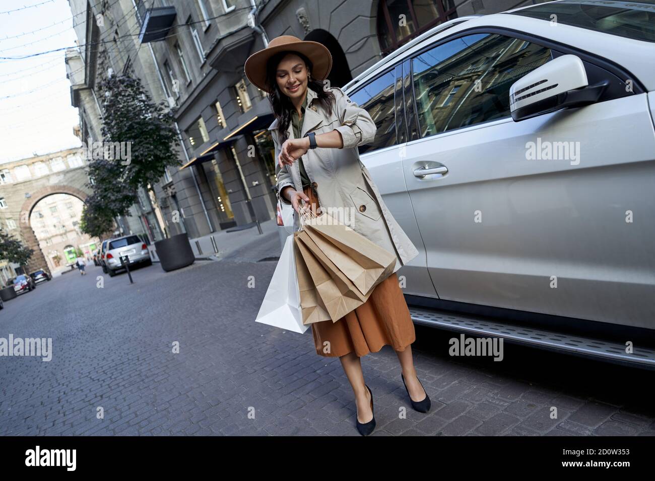 Junge modische Geschäftsfrau in Hut mit Papier Einkaufstaschen Überprüfung der Zeit auf ihrer Uhr, während sie auf der Stadtstraße in der Nähe von weißen Luxus-Auto. Einkaufen, Mode, Menschen Lebensstil Stockfoto