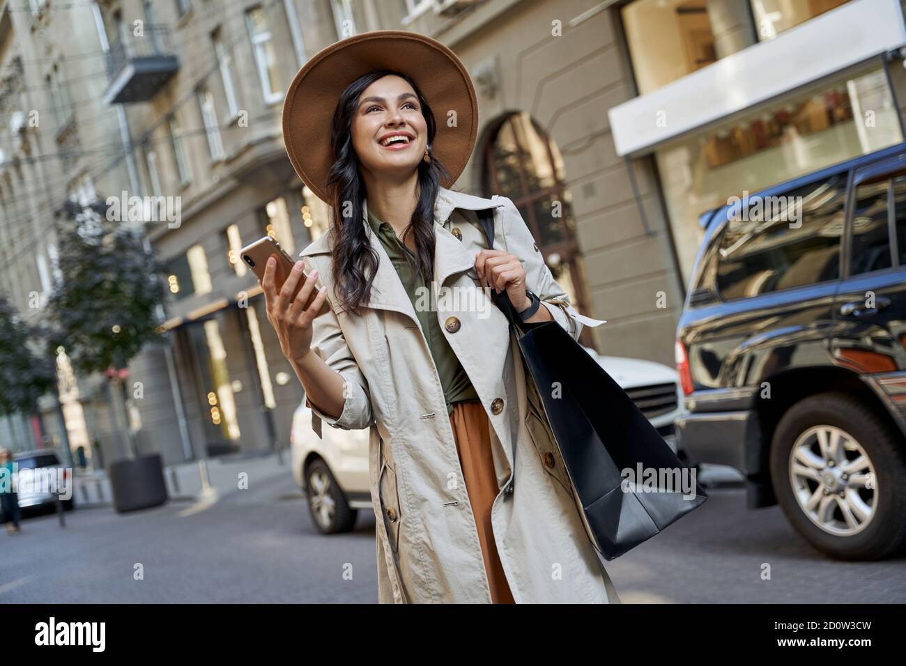 Gute Gefühle nach dem Einkaufen. Junge schöne stilvolle Frau in Hut mit Einkaufstasche hält ihr Smartphone, Blick zur Seite und lächelnd beim Gehen auf den Straßen der Stadt. Mode, Lifestyle Stockfoto
