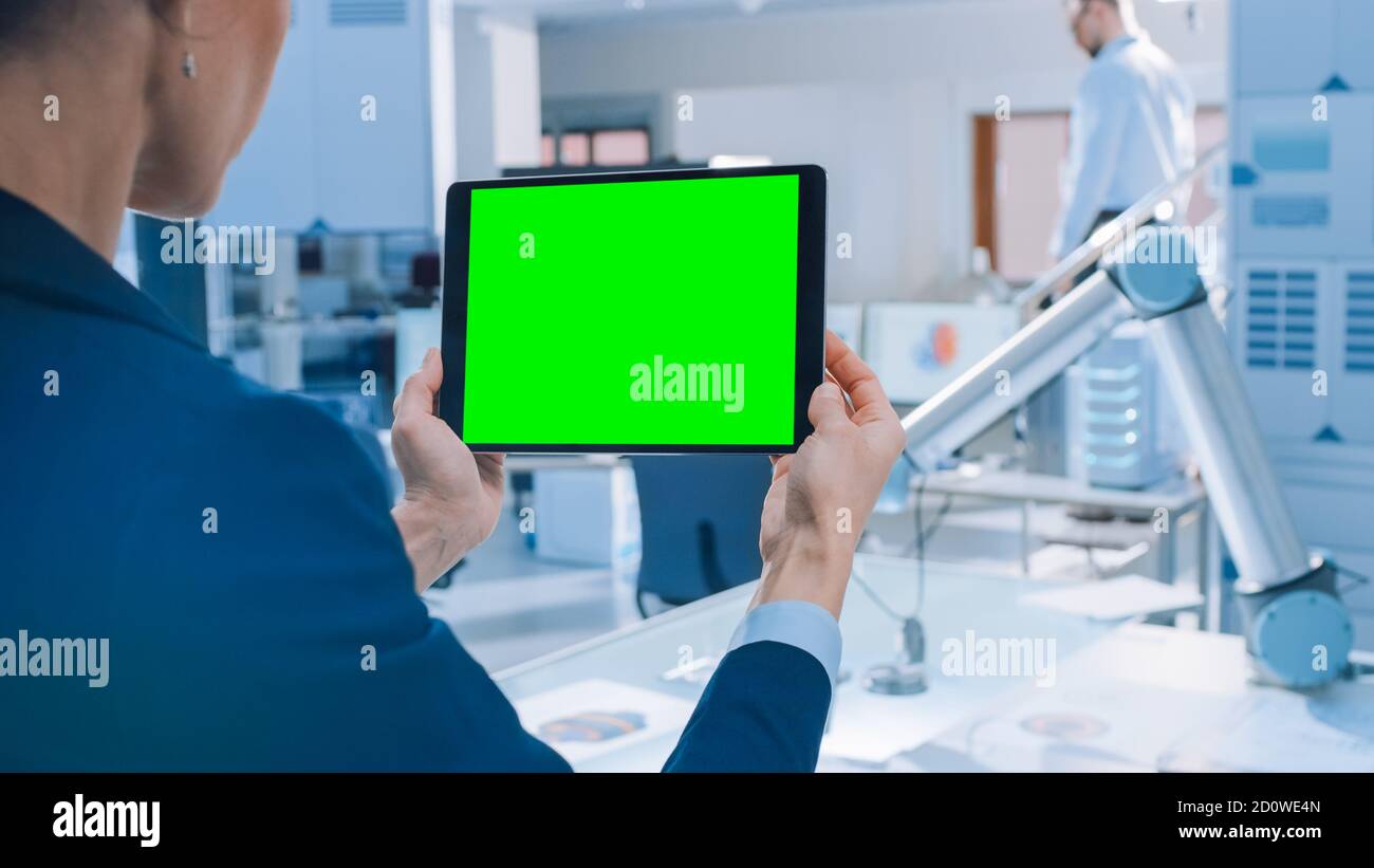 Der Techniker hält den digitalen Tablet-Computer mit dem grünen Mock-up-Bildschirm. Im Hintergrund Team von Industrieingenieuren arbeiten mit funktionalen Roboter Arm in Stockfoto