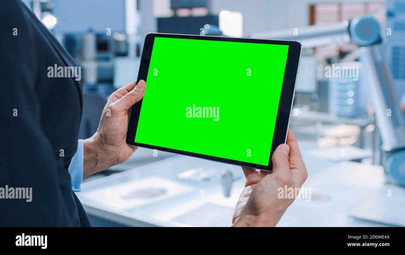 Der Techniker hält den digitalen Tablet-Computer mit dem grünen Mock-up-Bildschirm. Im Hintergrund Team von Industrieingenieuren arbeiten mit funktionalen Roboter Arm in Stockfoto