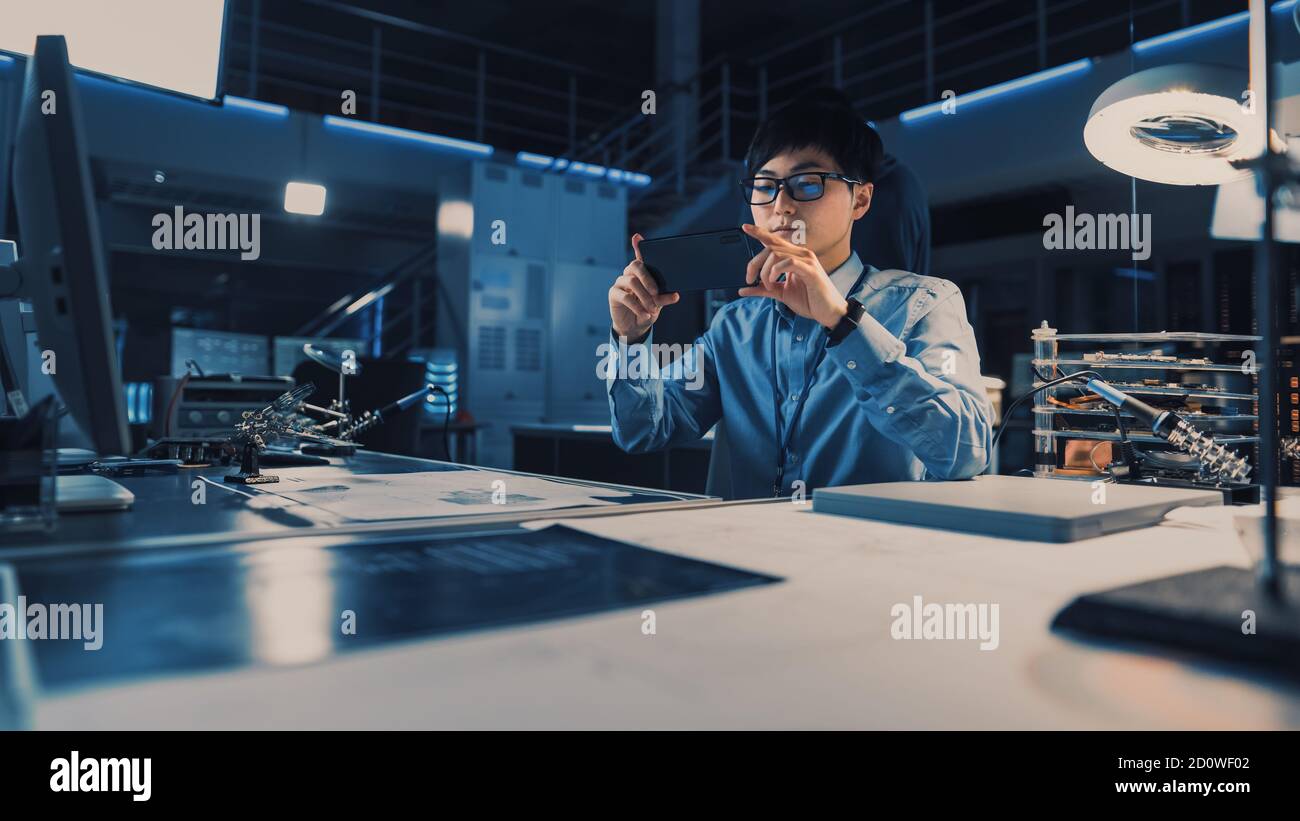 Professioneller japanischer Entwicklungsingenieur in blauem Hemd, der ein Foto nimmt Der technischen Zeichnungen auf seinem Smartpgone in der High Tech Forschungslabor Stockfoto