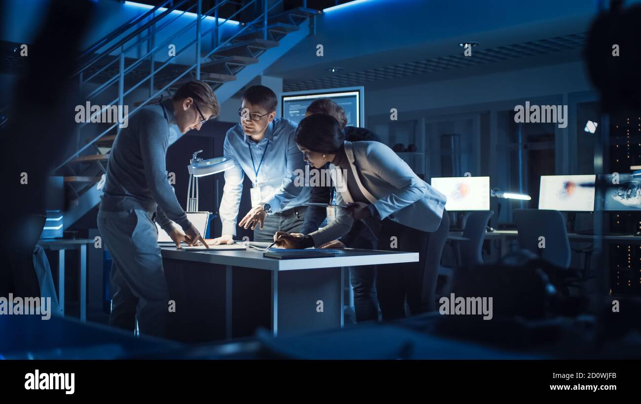 Vielfältiges Team von Elektronikentwicklern, die am Schreibtisch stehen und mit Dokumenten arbeiten, um Projektprobleme spät in der Nacht zu lösen. Spezialisten Stockfoto