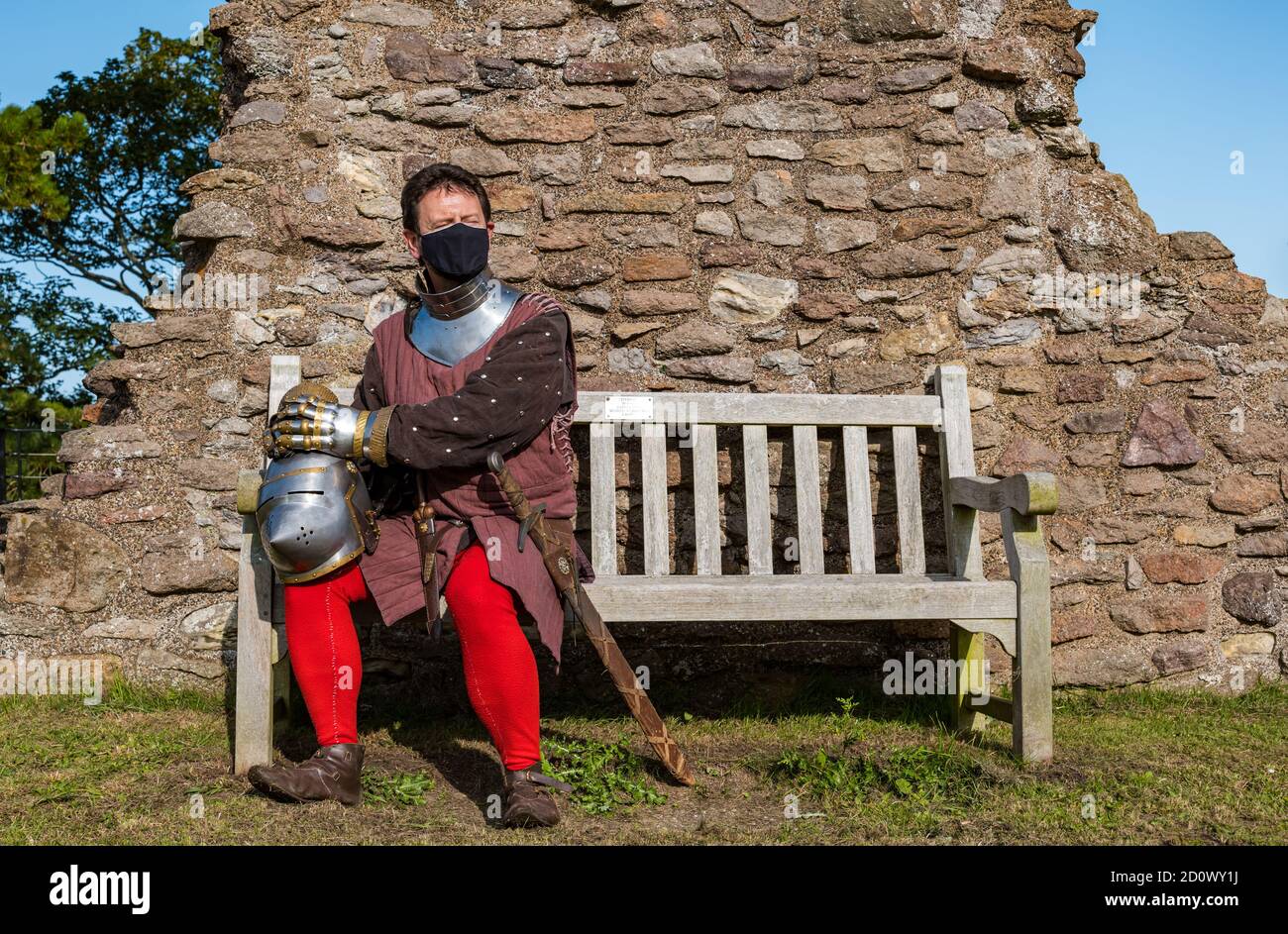 Mann mit Gesichtsmaske und einem Ritterkostüm mit Schwert und Helm sitzt auf der Bank in Dirleton Castle, East Lothian, Schottland, Großbritannien Stockfoto