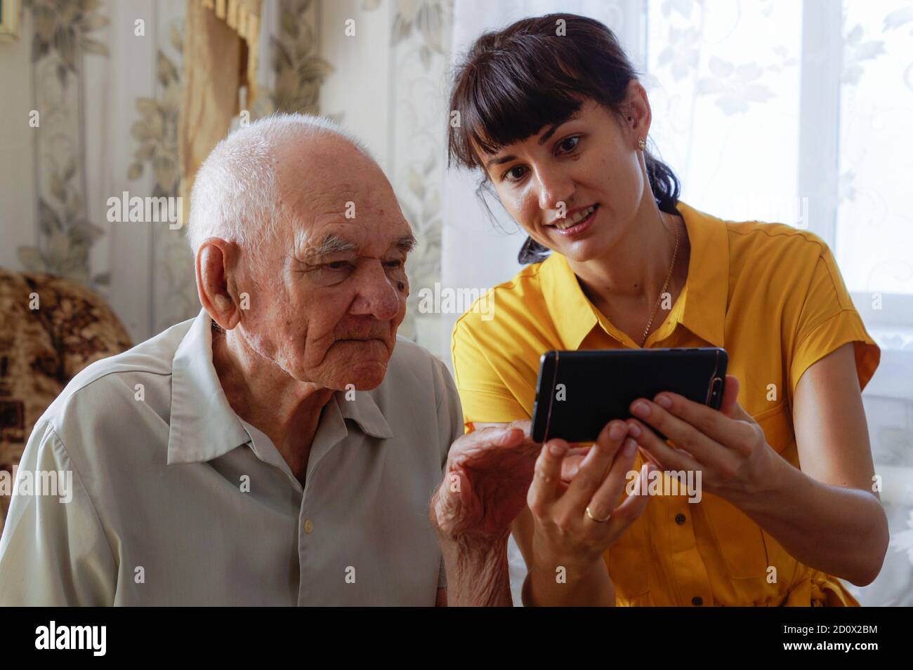 Ein Mann der 90er Jahre mit einer erwachsenen Enkelin, zusammen auf einem gemütlichen Sofa mit einem Telefon in den Händen sitzend, Blick auf Fotos. Eine Frau in einem gelben Kleid Stockfoto