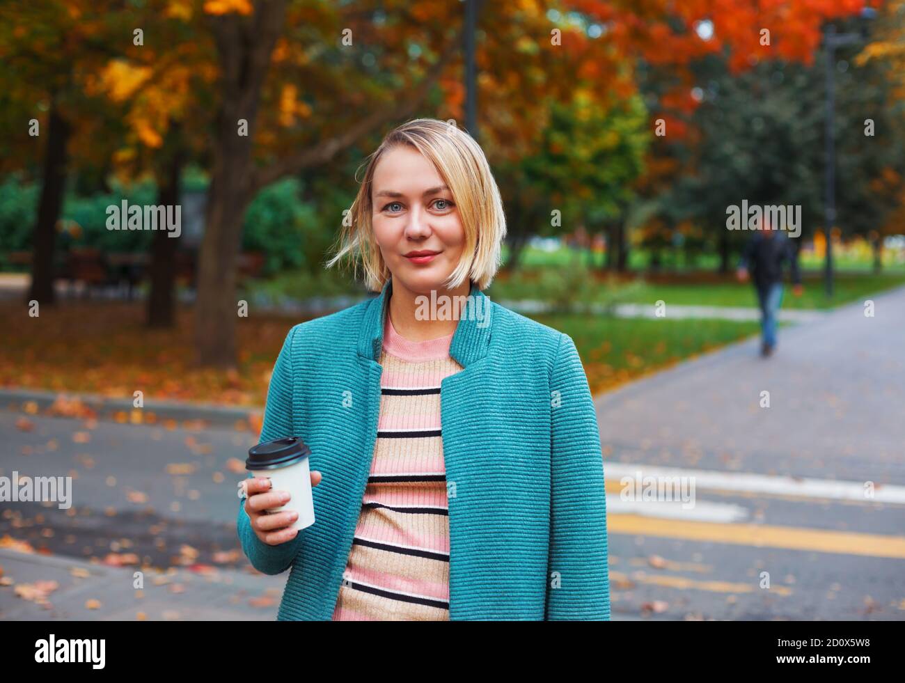 Lächelnde blonde kaukasische Frau mit Take Away Kaffeetasse zu Fuß im Herbst Park. Attraktive Frau, die auf einer Kreuzung steht Stockfoto