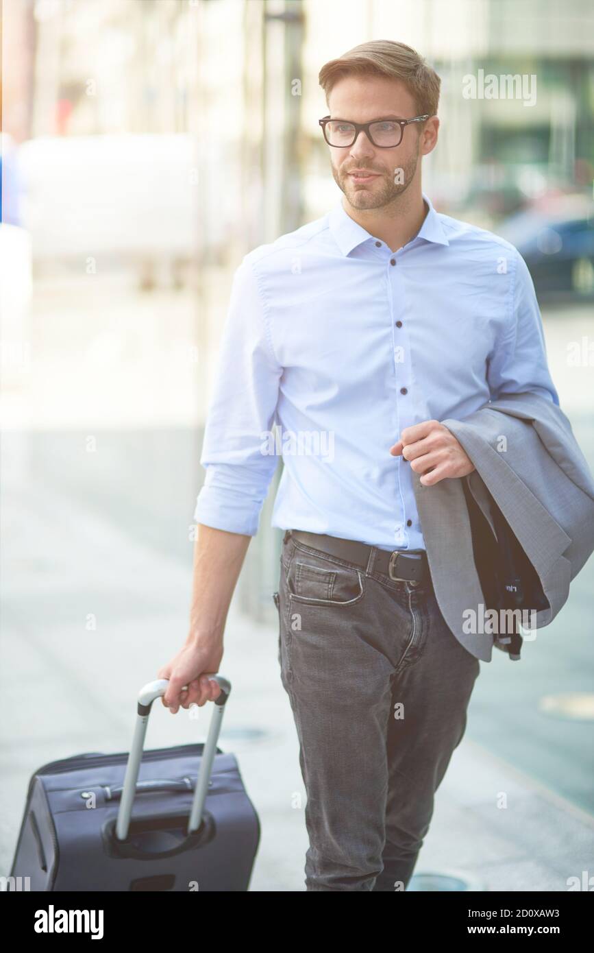 Vertikale Aufnahme eines jungen selbstbewussten Geschäftsmannes, der seinen Koffer zieht Und wegschauen, während man durch die Straßen der Stadt läuft Stockfoto