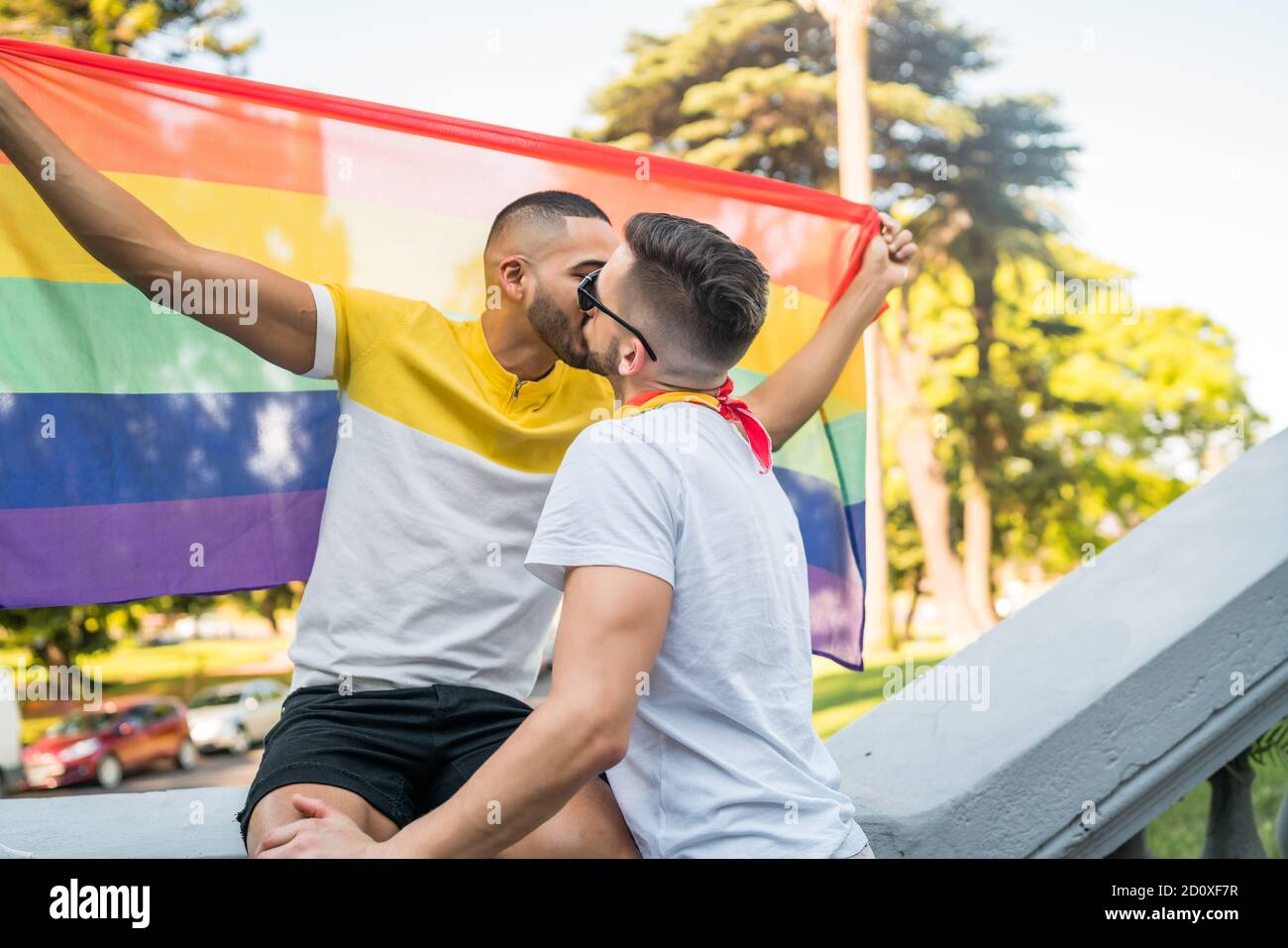 Porträt des jungen schwulen Paares, das sich küsst und seine Liebe zur Regenbogenfahne im Stret zeigt. LGBT und Love Konzept. Stockfoto