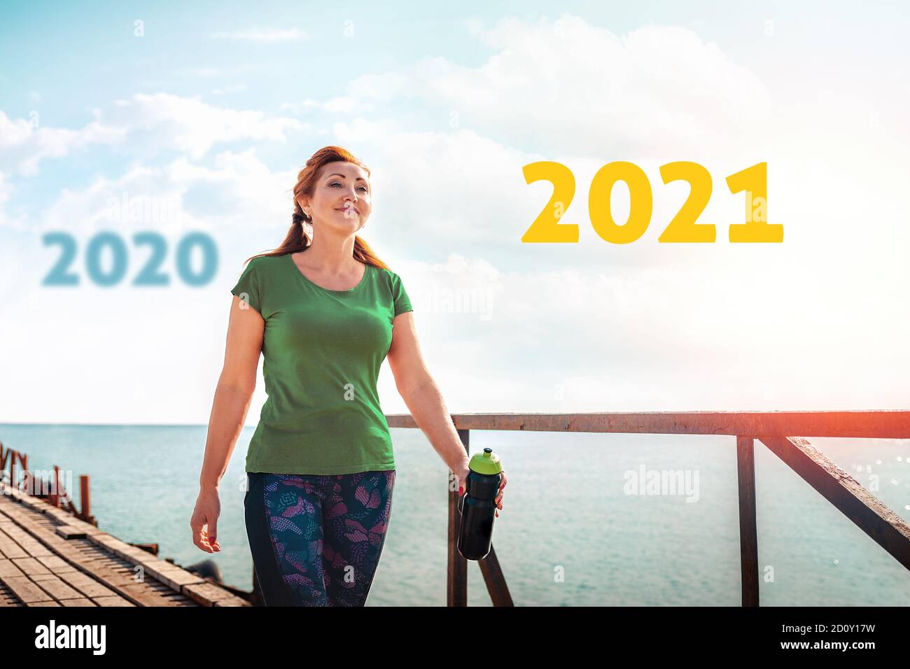 Jahr 2021, Kalender. Erwachsene glückliche Frau in Sportkleidung mit einem Shaker in den Händen, zu Fuß entlang der Pier. Im Hintergrund das Meer und der Himmel mit t Stockfoto