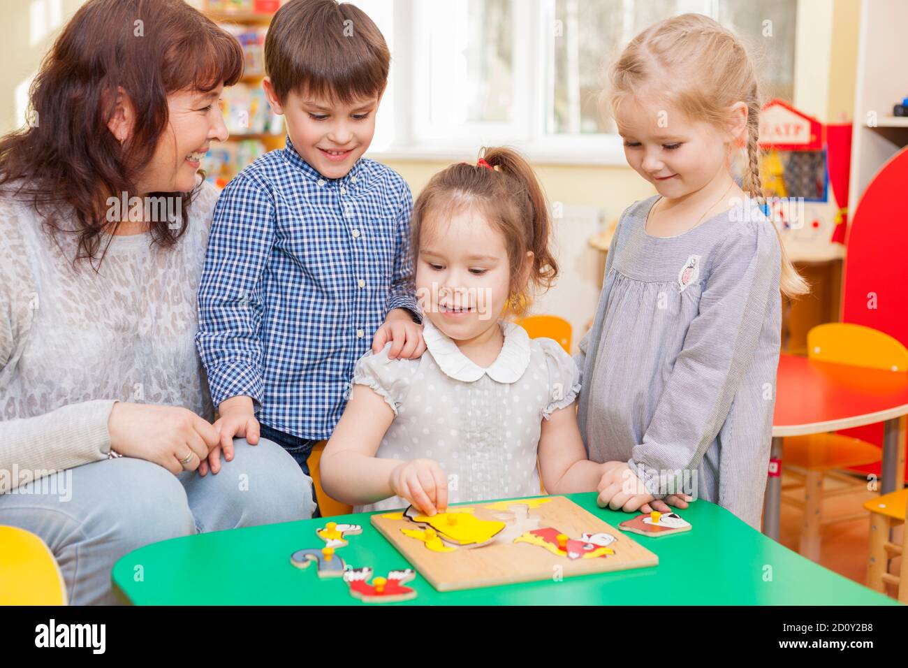 Hübsche kaukasische Mädchen, Kindergarten-Schüler, sammeln Puzzle im Klassenzimmer am grünen Tisch. Der Lehrer und zwei Schüler schauten zu und lächelten. Horizont Stockfoto