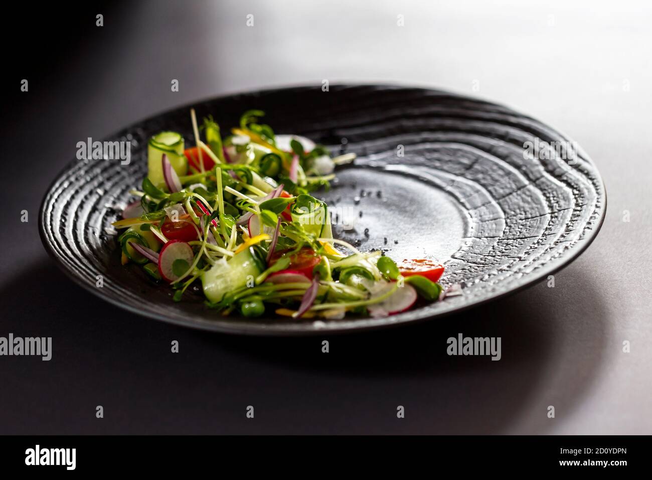 Authentischer frischer Gemüsesalat mit Sprossen und Erbsen schwarzer Pfeffer auf schwarzem Teller. Stimmungsvolle Morgenbeleuchtung, modischer, trendiger Spot-Weichfokus. Stockfoto