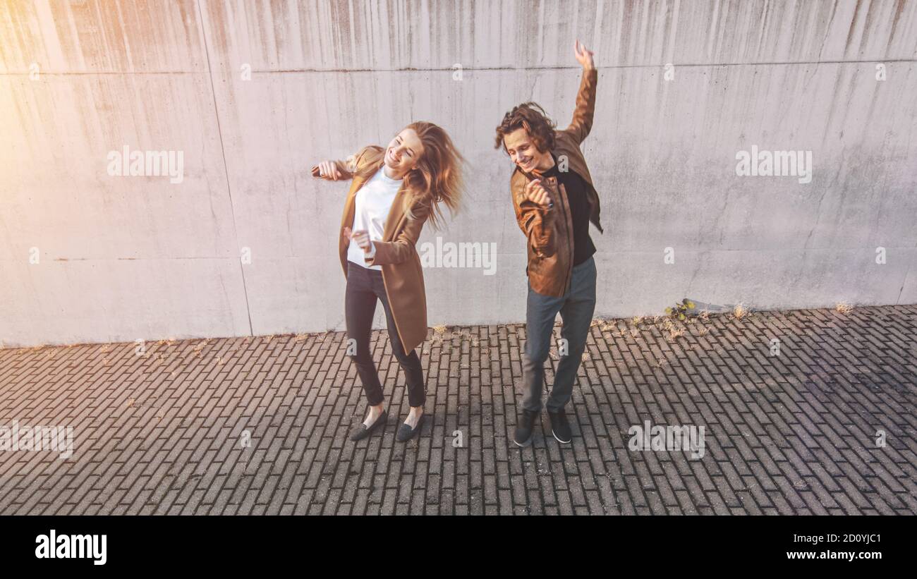 Fröhliches Mädchen und glücklicher junger Mann mit langen Haaren tanzen aktiv auf einer Straße neben einer städtischen Betonmauer. Sie tragen braune Lederjacke und Stockfoto