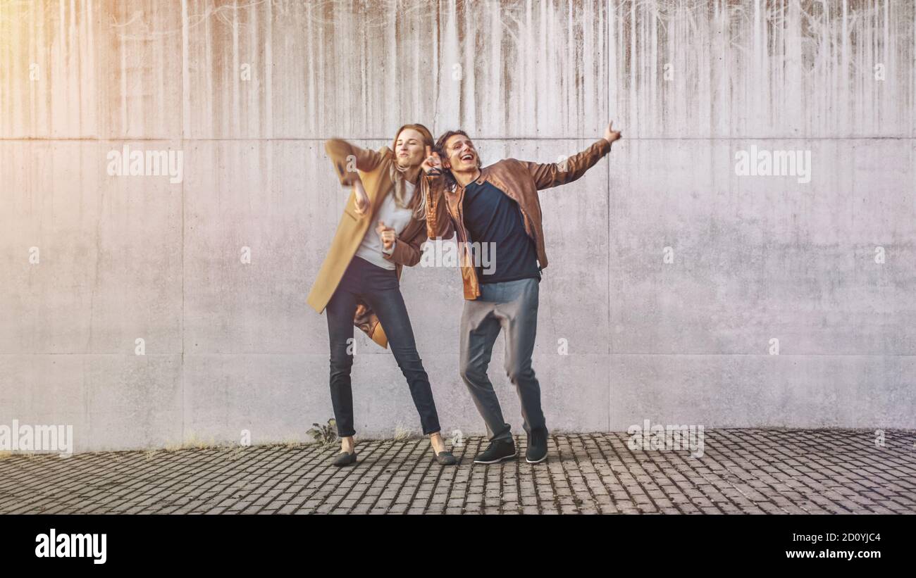 Fröhliches Mädchen und glücklicher junger Mann mit langen Haaren tanzen aktiv auf einer Straße neben einer städtischen Betonmauer. Sie tragen braune Lederjacke und Stockfoto