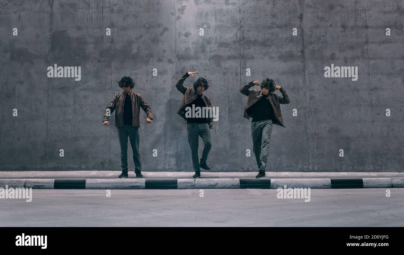 Multiple Exposure Shot eines coolen geklonten jungen Hipstermannes mit langen Haaren steht in Tanzposen auf einer Straße neben einer großen Betonwand. Er trägt einen Stockfoto