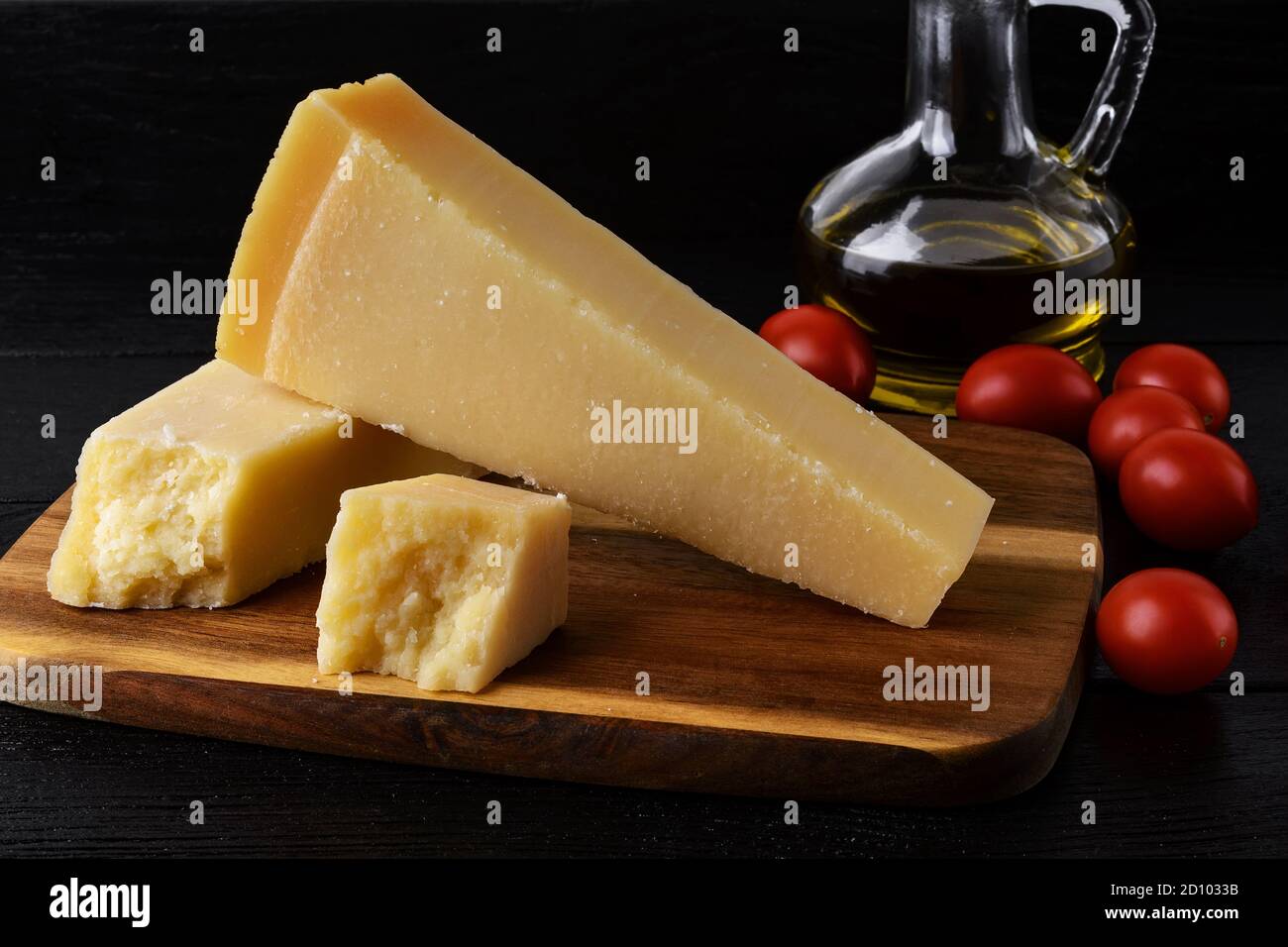 Italienischer Hartkäse Grana Padano oder Parmesan auf einem braunen Holzschnitt-Boad, kleine rote Tomaten und Olivenöl auf schwarzem Holztisch. Low-Key-Bild. Stockfoto